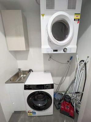 A compact laundry room with a front-loading washing machine, a stainless steel sink, and a vacuum cleaner. - New L63 Hill River CBD View&Pool & Sauna (Surfers Paradise)