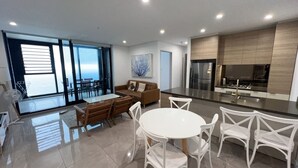 A modern kitchen with a central island, dining area with white chairs, and a large window providing a view. - Meriton L47 with SeaViews and CarPark (Surfers Paradise)