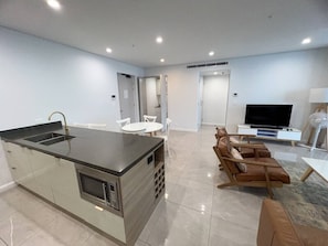 A modern kitchen with a central island, a dining area with chairs, a sofa, a flat-screen TV, and a fan. - Meriton L47 with SeaViews and CarPark (Surfers Paradise)