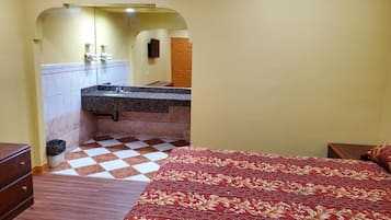 A hotel room with a checkered floor, a bed with a patterned bedspread, a wooden nightstand, and a bathroom with a tiled wall and a sink.