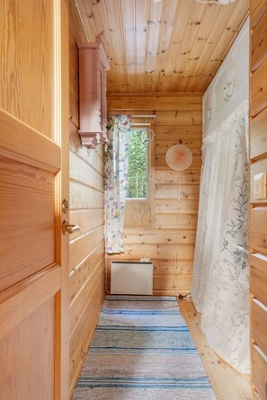 A narrow wooden hallway with a door on the left, a window with floral curtains, a radiator, and a patterned rug.