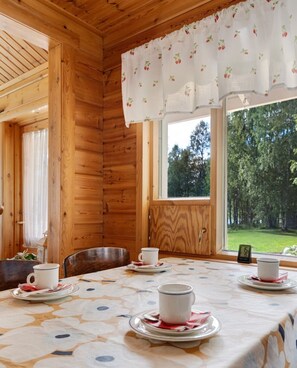 A wooden cabin interior with a dining table set for tea, a window with a view of a forest, and a clock on the wall.