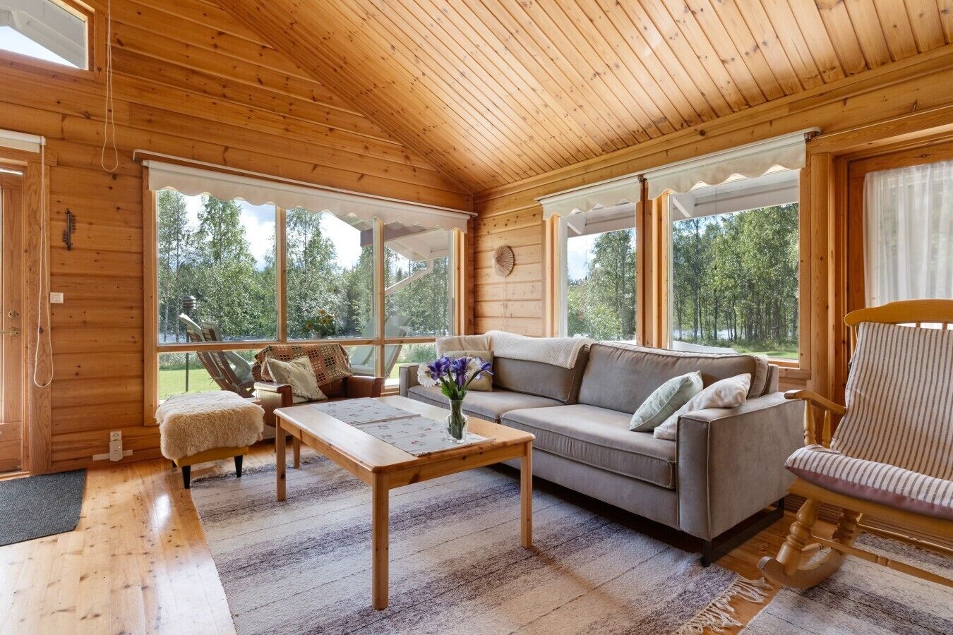 A cozy cabin interior with a wooden ceiling, large windows, a sofa, a chair, and a coffee table.