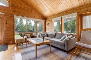 L’intérieur d’un chalet accueillant, avec un plafond en bois, de grandes fenêtres, un canapé, un fauteuil et une table basse.