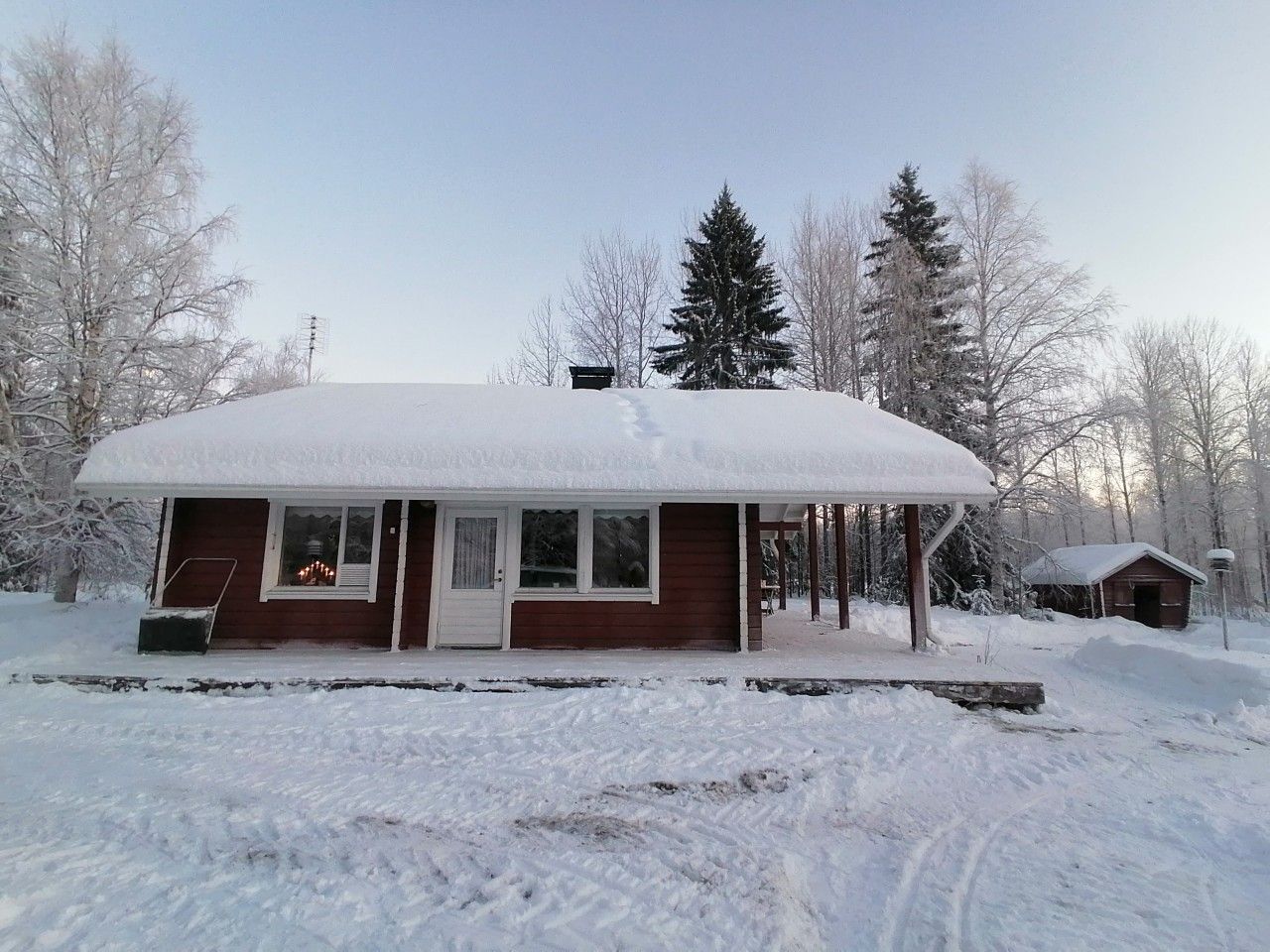 A snow-covered cabin with a porch, a small attached shed, and a clear sky.