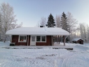 Une cabane recouverte de neige, dotée d’un porche, d’une petite remise attenante et surplombée par un ciel dégagé.