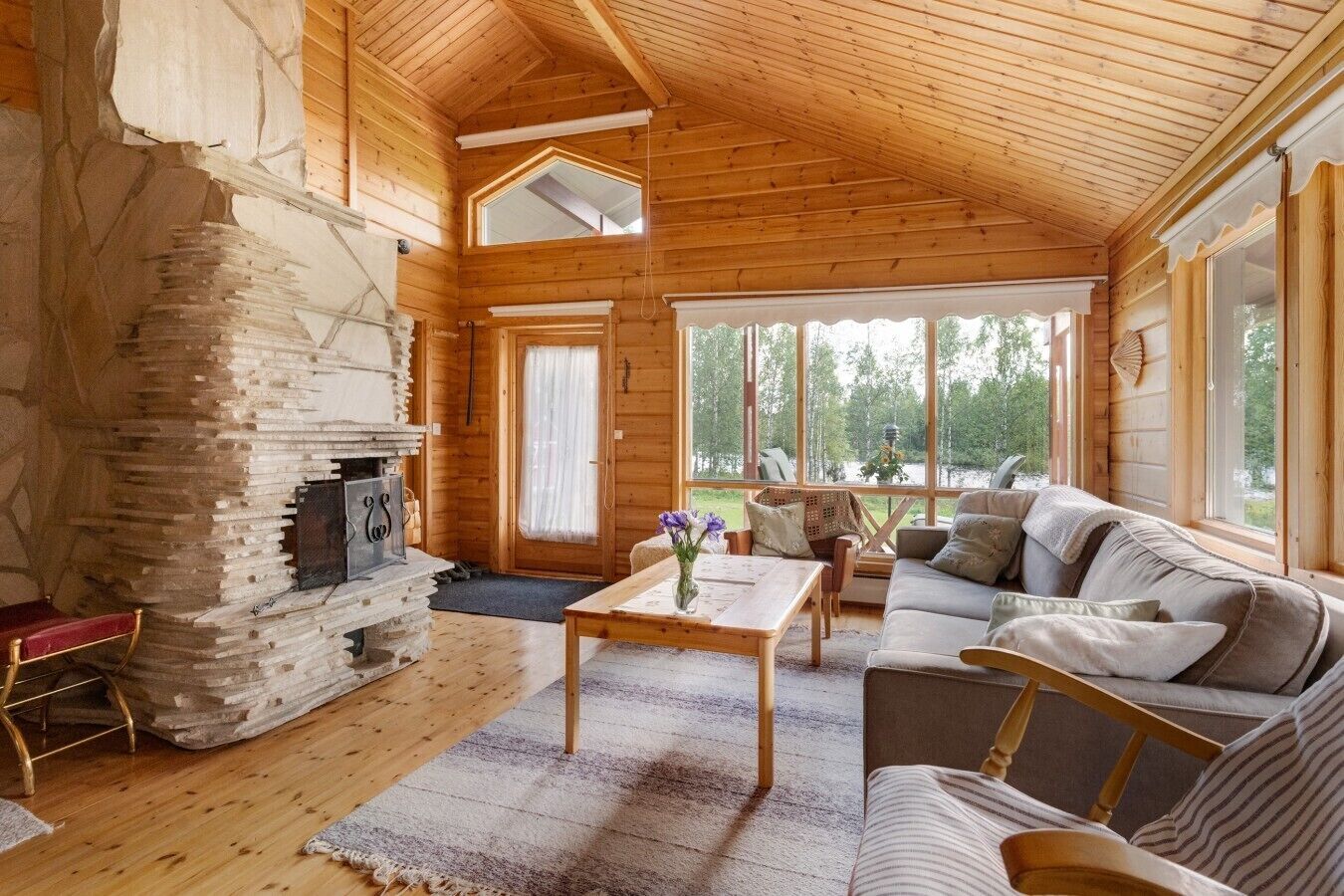 A cozy cabin interior with a stone fireplace, a sofa, a coffee table, and large windows offering a view of the outdoors.