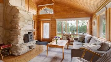 A cozy cabin interior with a stone fireplace, a sofa, a coffee table, and large windows offering a view of the outdoors.