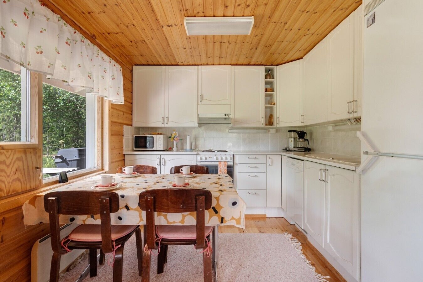 Cocina compacta con alacenas blancas, microondas, estufa y cafetera. Hay una mesa de comedor con sillas y una ventana con vistas a zonas verdes.