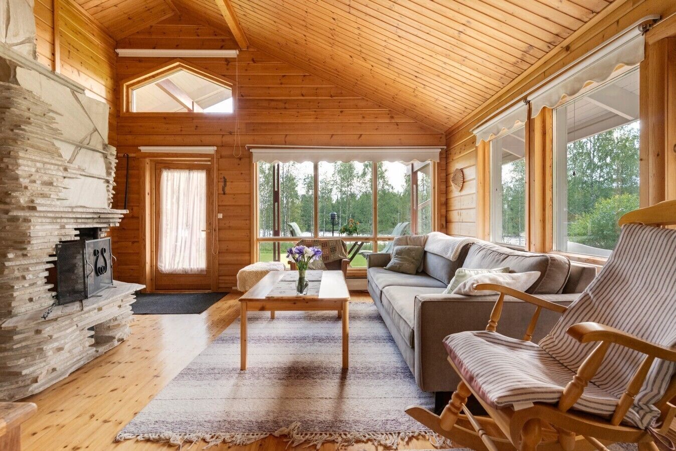 Interior de una cabaña de madera con chimenea, sofá, mecedora y ventanas amplias.
