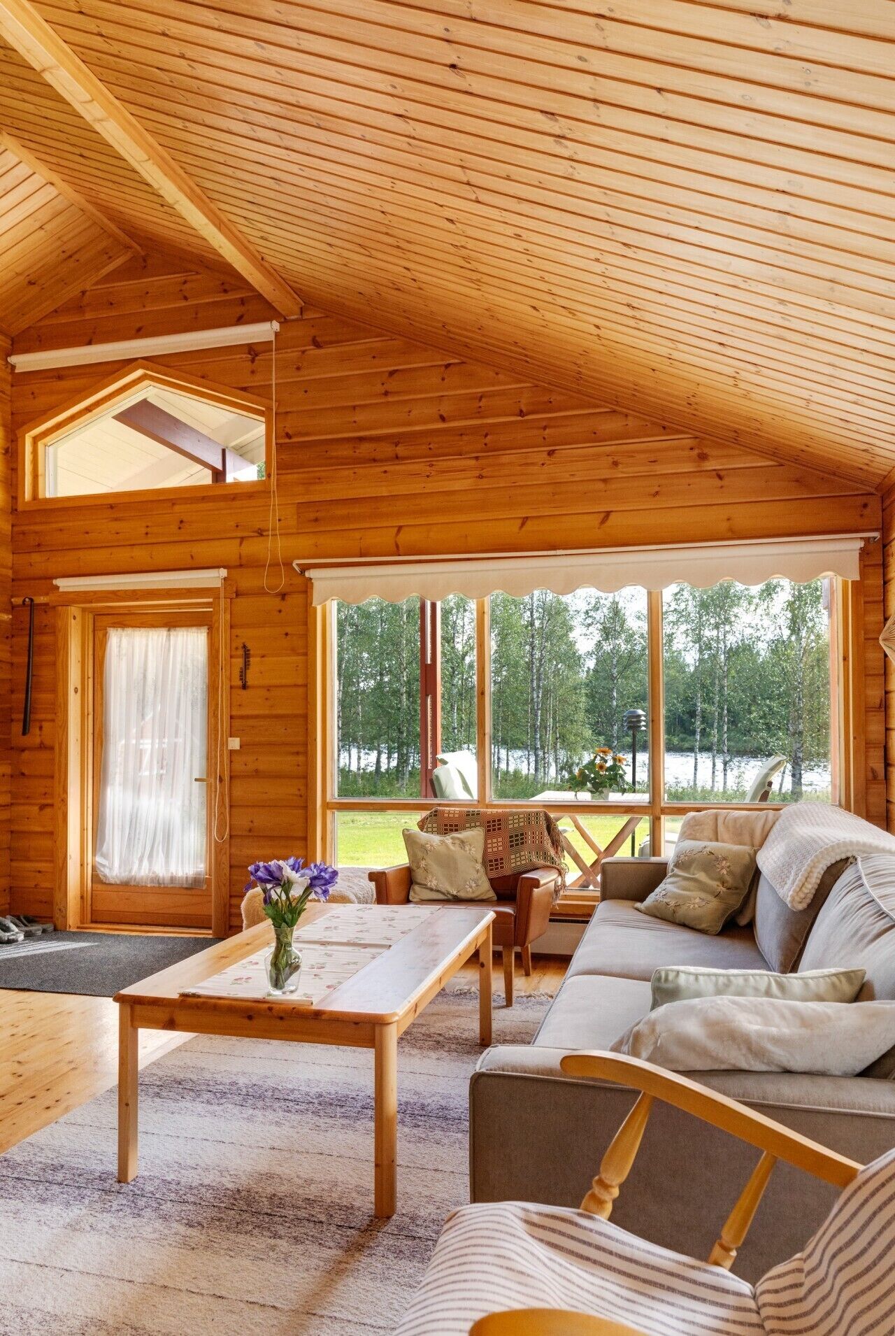 A wooden cabin interior with a sofa, a table, and a window overlooking a forest.