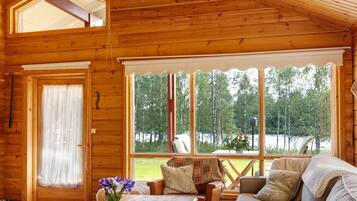 A wooden cabin interior with a sofa, a table, and a window overlooking a forest.
