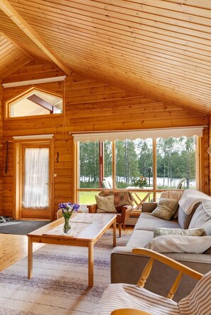 A wooden cabin interior with a sofa, a table, and a window overlooking a forest.