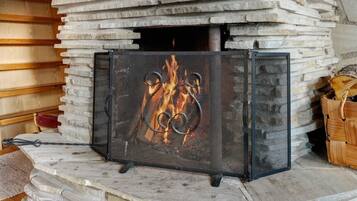 Un camino in pietra con un paravento metallico, circondato da tronchi di legno.
