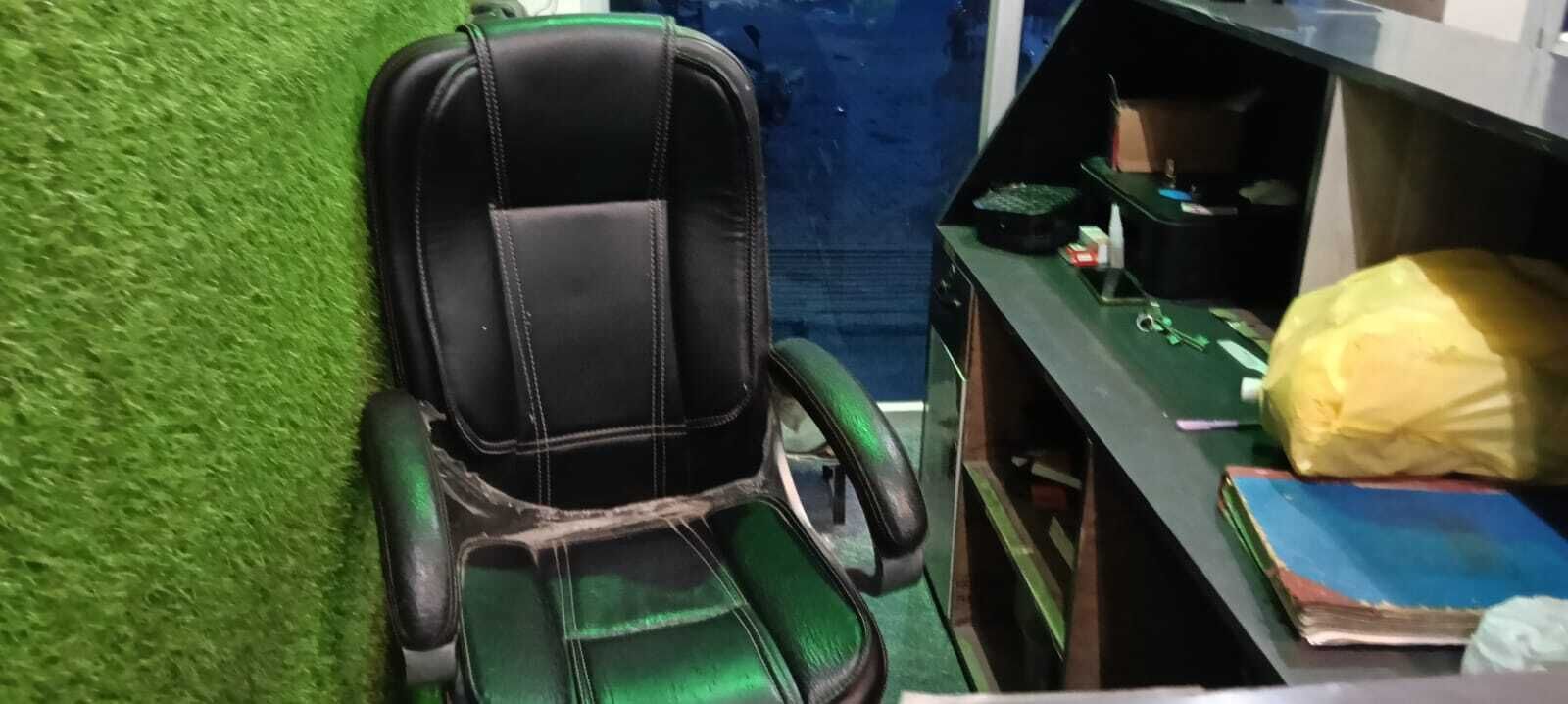 Um quarto com parede de grama sintética verde, uma cadeira de escritório preta e verde, uma mesa com uma bolsa amarela e um livro azul.