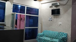 A room with a sofa, a television mounted on the wall, and a window with a view. - The Singh Hotel (Auraiya)