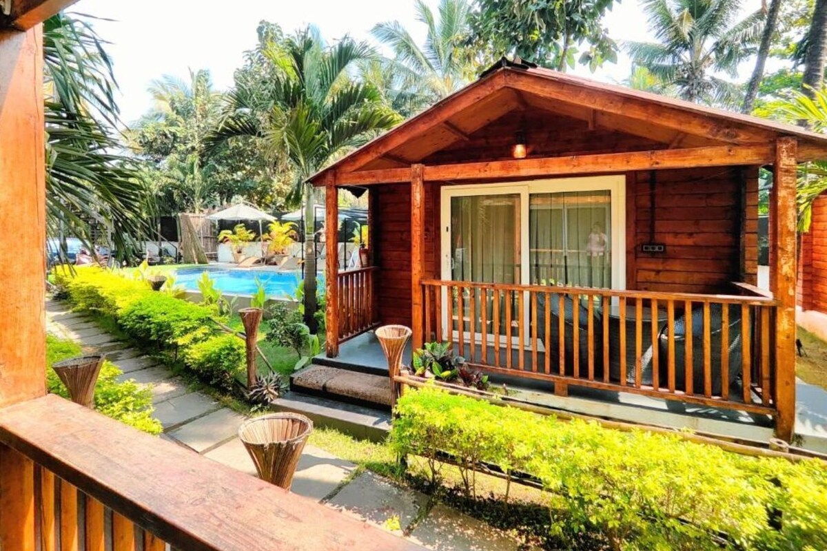 Una cabaña de madera con balcón, rodeada de exuberante vegetación y una piscina.