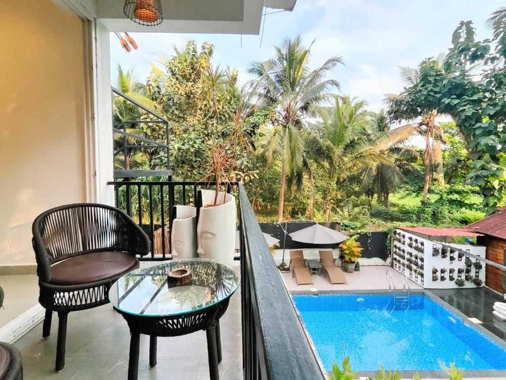 A balcony with a glass table, a chair, and a pool area with lounge chairs and umbrellas.