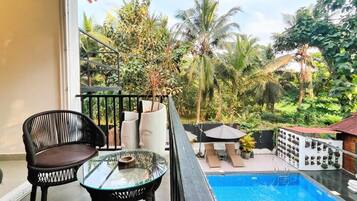 A balcony with a glass table, a chair, and a pool area with lounge chairs and umbrellas.