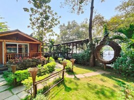 Una cabaña de madera con un porche cubierto, rodeada de abundante vegetación y una estructura circular tejida.