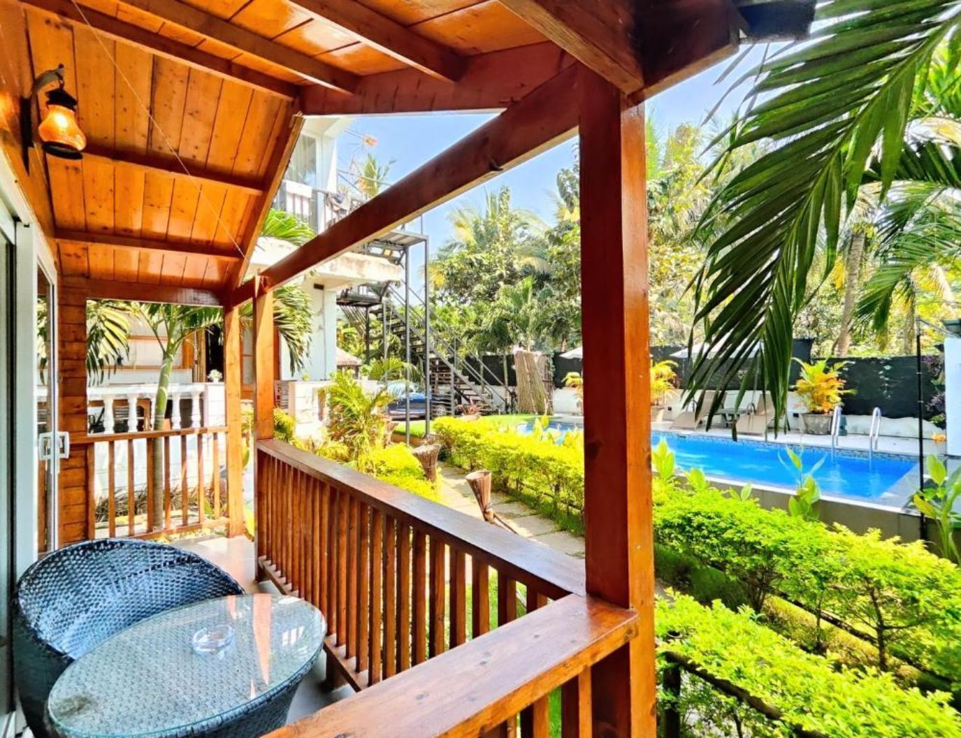 Terraza de madera con mesa y sillas de vidrio, con vista a una piscina y exuberante vegetación.