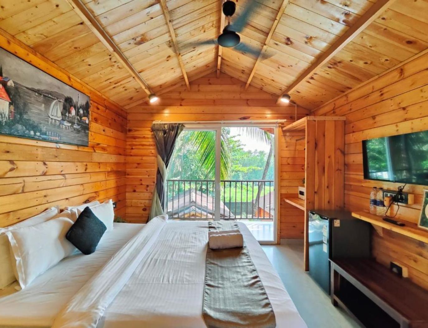 A wooden cabin room with a bed, a TV, and a balcony with a view of greenery.