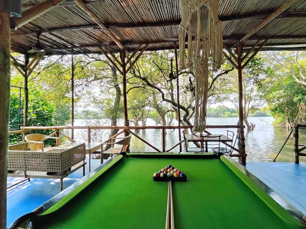 Een pooltafel met een groene viltkleed, omringd door een houten terras met rieten dak, met uitzicht op een waterpartij.