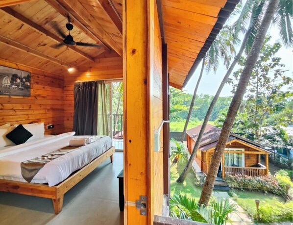 A wooden cabin with a bed, a ceiling fan, and a view of a tropical garden. - Trideus Riviera - Riverside Resort, Candolim Goa (Candolim)