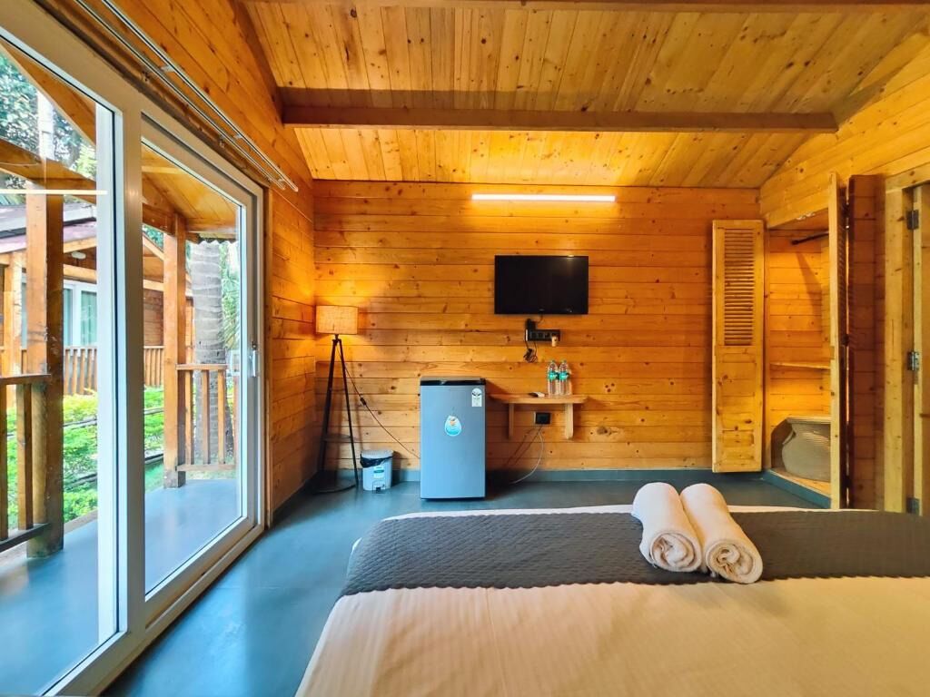 Quarto de cabana de madeira com uma cama, televisão, pequeno frigorífico e janela.
