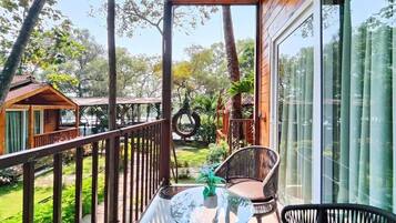 A balcony with a glass table and two wicker chairs, overlooking a garden with trees and a wooden structure in the background.