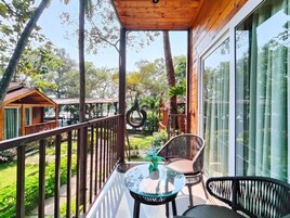 Uma varanda com mesa de vidro e duas cadeiras de vime, com vista para um jardim com árvores e uma estrutura de madeira ao fundo.