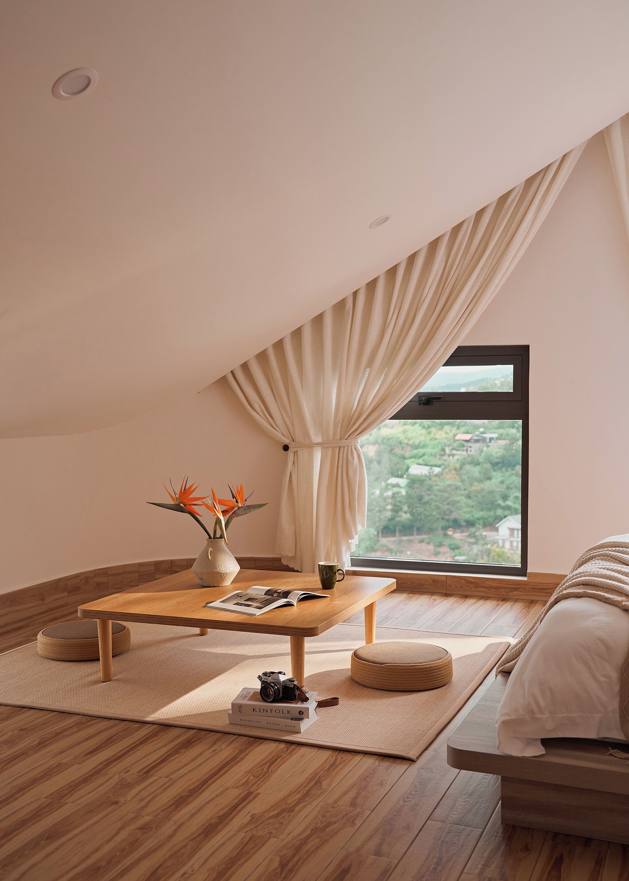 A cozy room with a wooden table, a window with curtains, a bed, and a view of greenery outside.