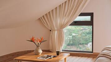 A cozy room with a wooden table, a window with curtains, a bed, and a view of greenery outside.