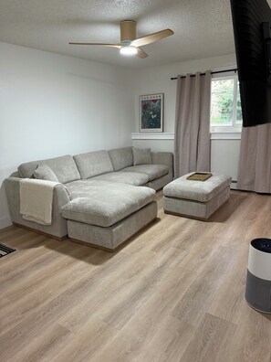 Ein modernes Wohnzimmer mit einem Ecksofa, einem Hocker, einem Deckenventilator und einem Fenster mit Vorhängen.