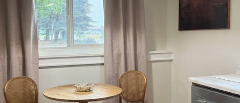 Un comedor con una mesa redonda, dos sillas de mimbre, una lámpara colgante, una ventana con cortinas y un cuadro en la pared.