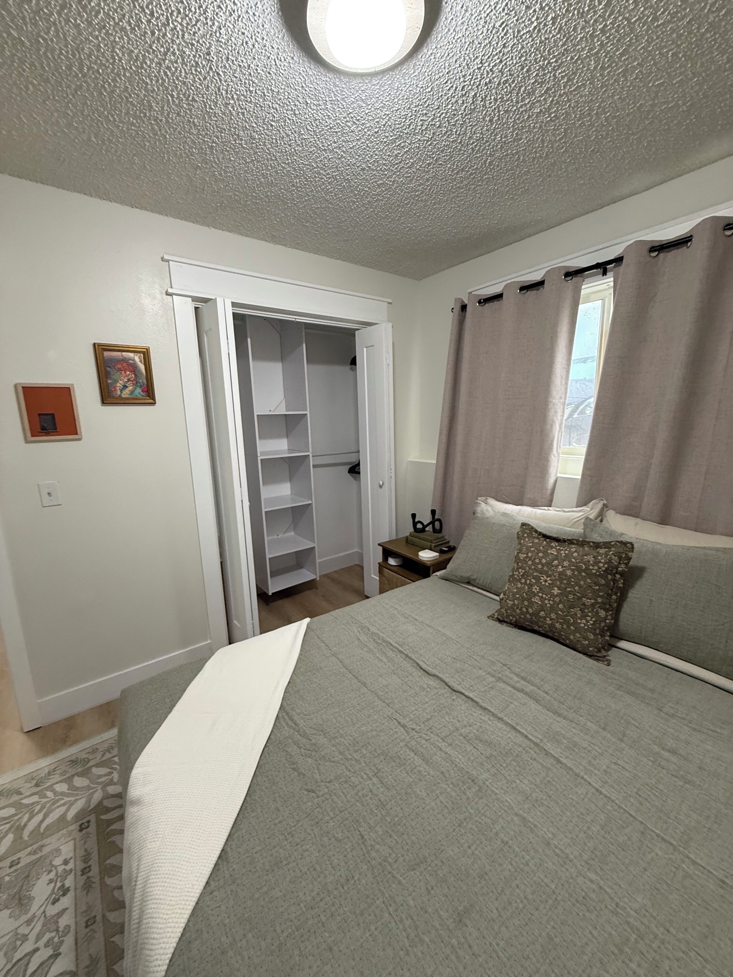 Un dormitorio con cama, mesita de noche, armario y ventana con cortinas.