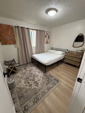 Ein Schlafzimmer mit einem Bett, einer Kommode, einem Spiegel, einem Fenster mit Vorhängen und einem gemusterten Teppich.