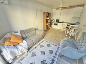 Ein Wohnzimmer mit Sofa, Sessel und einem gemusterten Teppich. Im Hintergrund ist eine Küche mit weißen Schränken und ein Essbereich mit Tisch und Stühlen zu sehen.