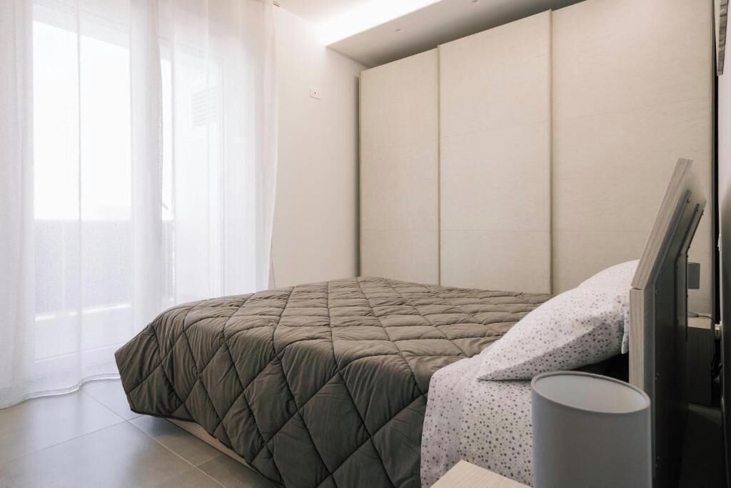 Ein Schlafzimmer mit einem Bett, einem Nachttisch und einem Fenster mit leichtem Vorhang.