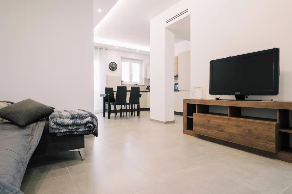 A modern living room with a flat-screen TV on a wooden stand, a sofa with a blanket, and a dining area with chairs and a table.