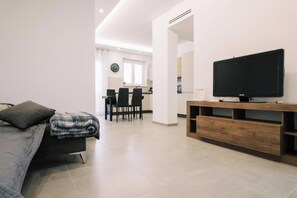 A modern living room with a flat-screen TV on a wooden stand, a sofa with a blanket, and a dining area with chairs and a table. - Daisy Apartment – Modern Comfort with a Balcony in Molfetta (Molfetta)