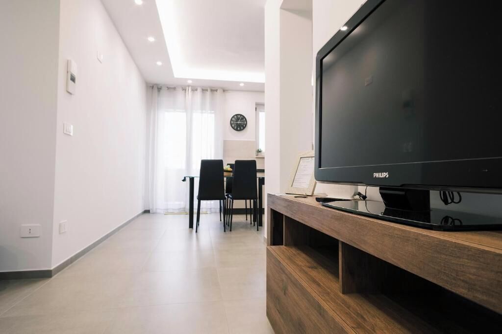 Ein modernes Wohnzimmer mit einem großen Flachbildfernseher auf einem Holzboden, einem Essbereich mit Stühlen und Tisch sowie einer Küche im Hintergrund.