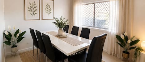 Un comedor con una mesa blanca, sillas negras, un jarrón con una planta y una ventana con cortinas.