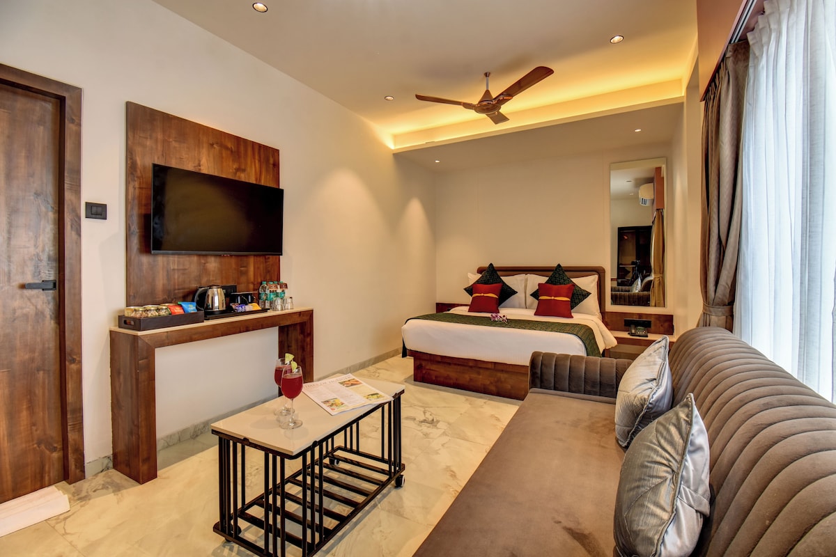 Une chambre d’hôtel avec un lit, un canapé, un téléviseur fixé au mur, un ventilateur de plafond et une petite table avec un verre à vin.