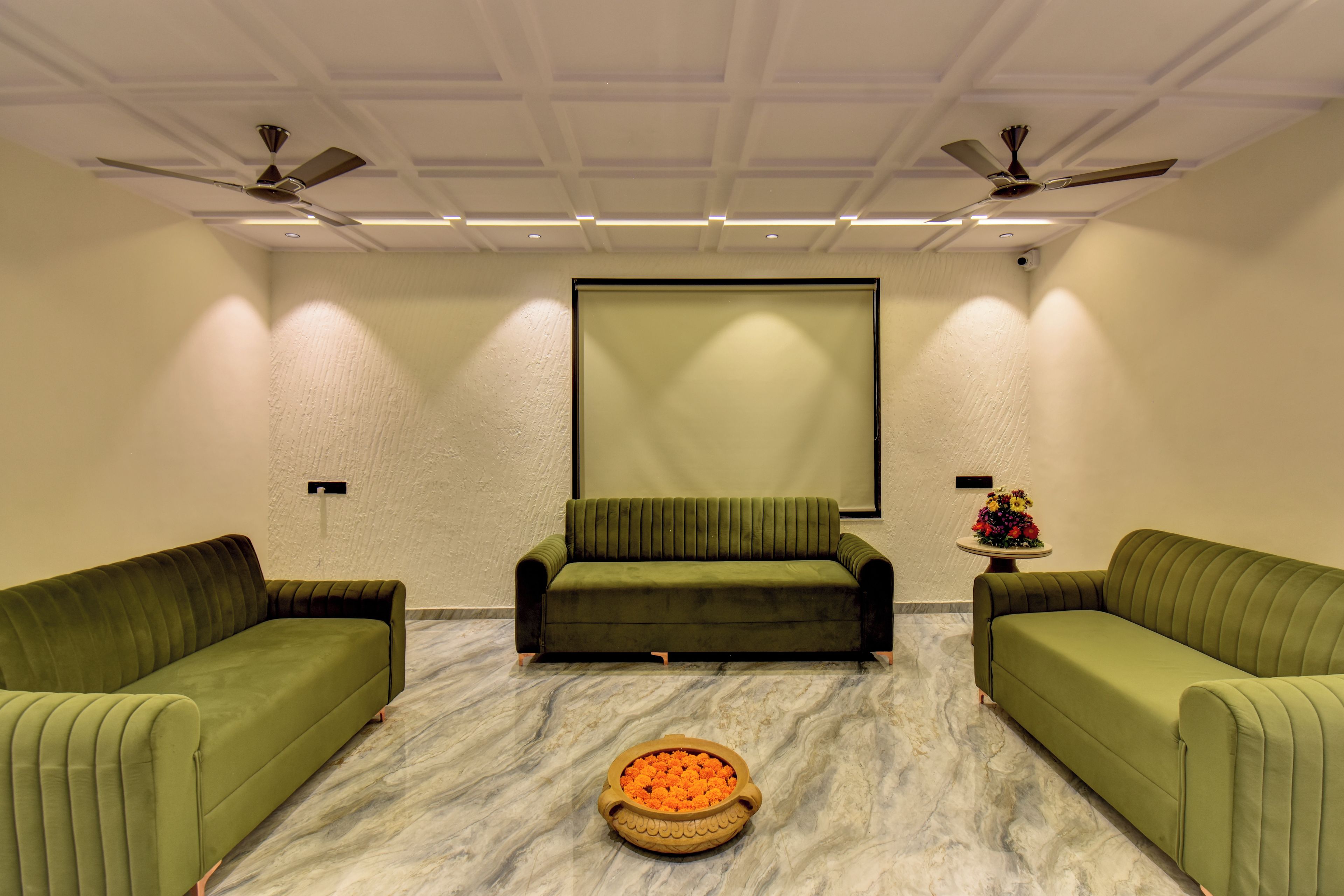 A room with two green sofas, a central green sofa, a marble floor, a ceiling fan, recessed lighting, and a large blank screen on the wall.