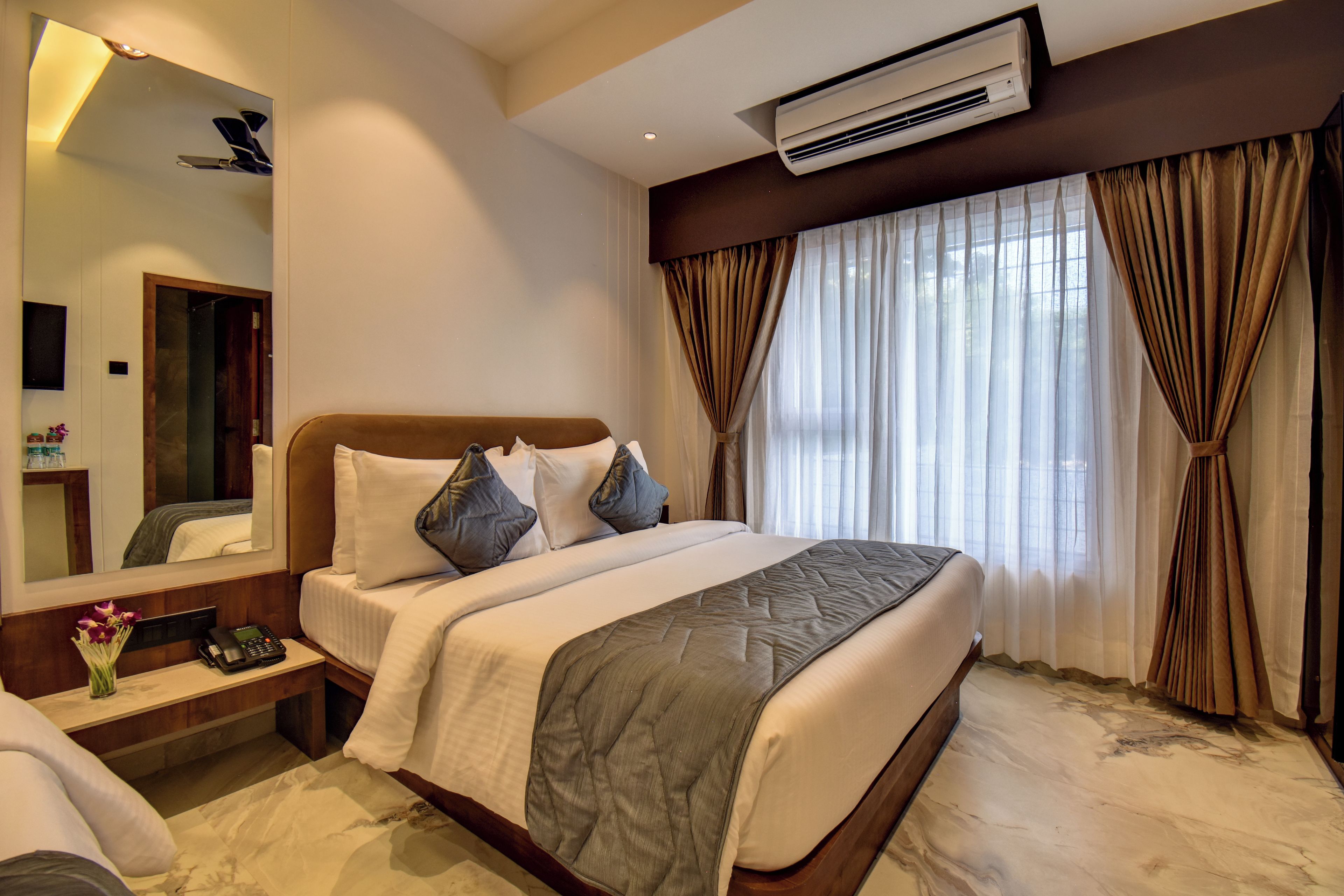 A hotel room with a bed, a bedside table with a phone, a mirror, and an air conditioning unit.