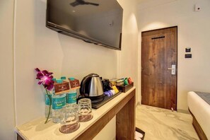 A hotel room with a flat-screen TV mounted on the wall, a wooden door, a bedside table with bottled water, a kettle, and a flower. - Hotel Orchid by Marigold, Shirdi - Premium Comfort Near Sai Baba Temple (Rahata)