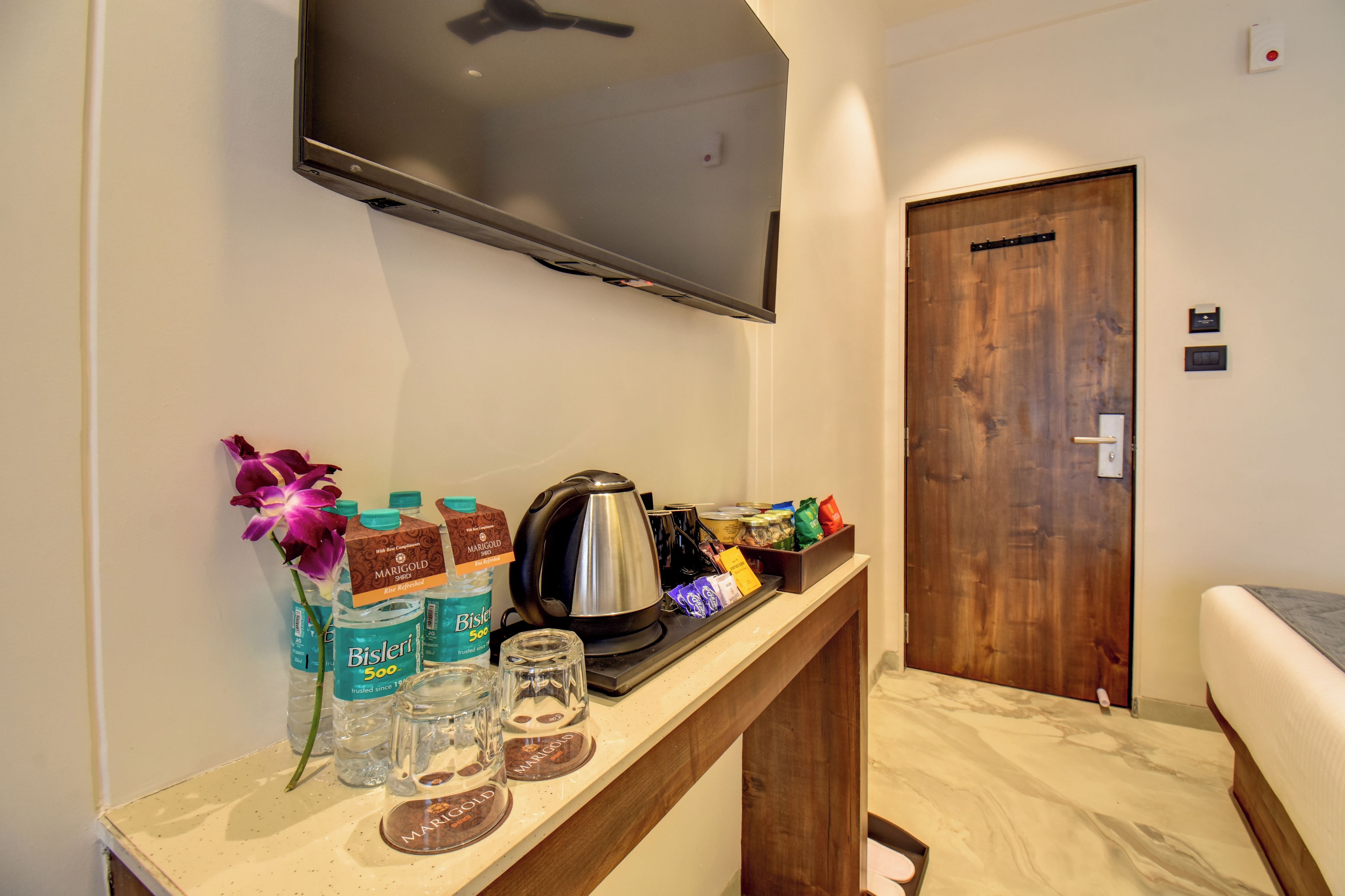 A hotel room with a flat-screen TV mounted on the wall, a wooden console table with a kettle, Bisleri water bottles, and a flower arrangement.