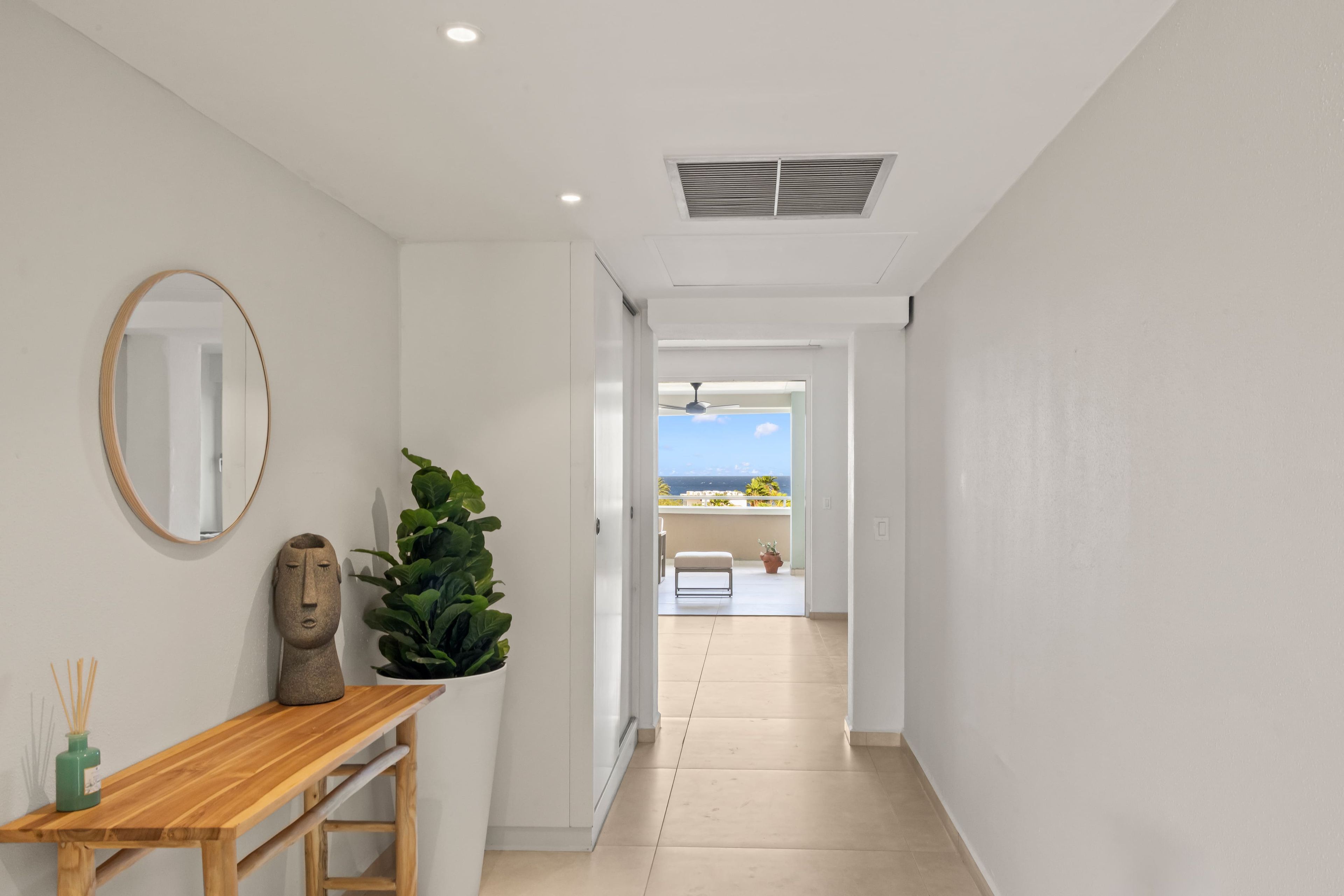 A hallway with a wooden bench, a mirror, and a potted plant. It leads to a balcony with a view of the sea.