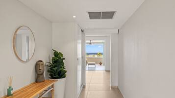 A hallway with a wooden bench, a mirror, and a potted plant. It leads to a balcony with a view of the sea.